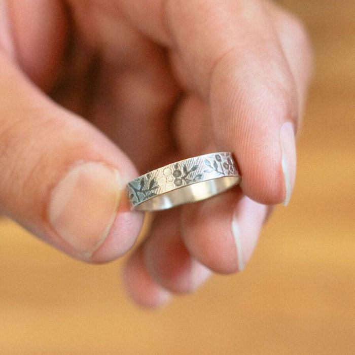 Silver ring with floral patterns held between fingers against a blurred background