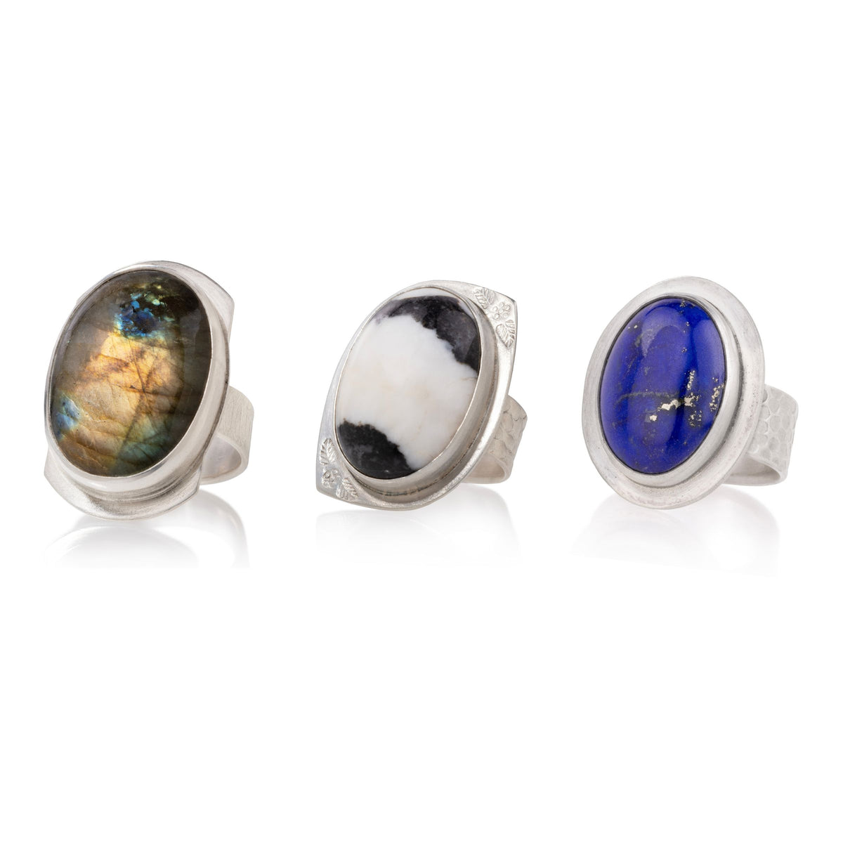 Three silver rings with large oval cabochon gemstones are displayed on a white reflective surface. The left ring features a brown and gold iridescent stone, the middle ring has a black and white marbled stone, and the right ring is a deep blue stone with small gold inclusions.