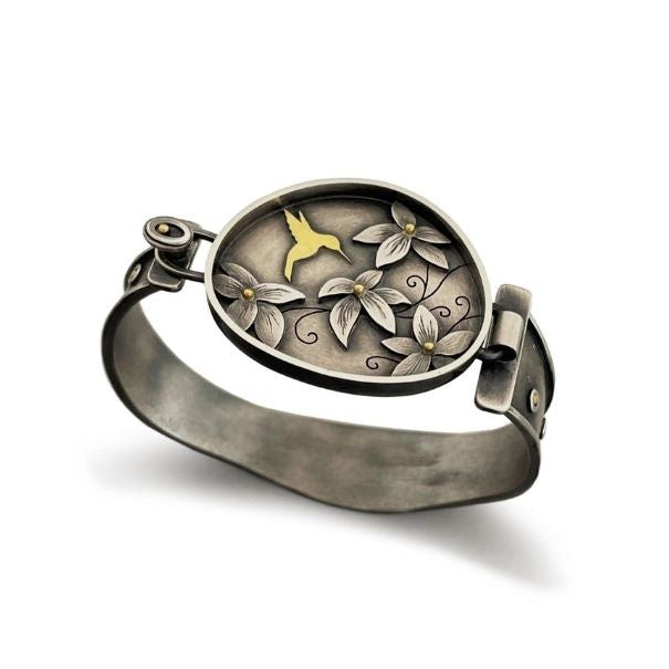 A silver-toned cuff bracelet with an oval hinged cover featuring a floral and hummingbird design in silver and gold, shown on a white background.