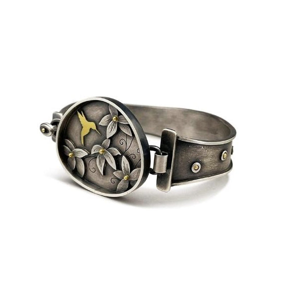 An oval silver-toned cuff bracelet featuring an openwork floral design with a small gold bird in the center, shown on a plain white background.