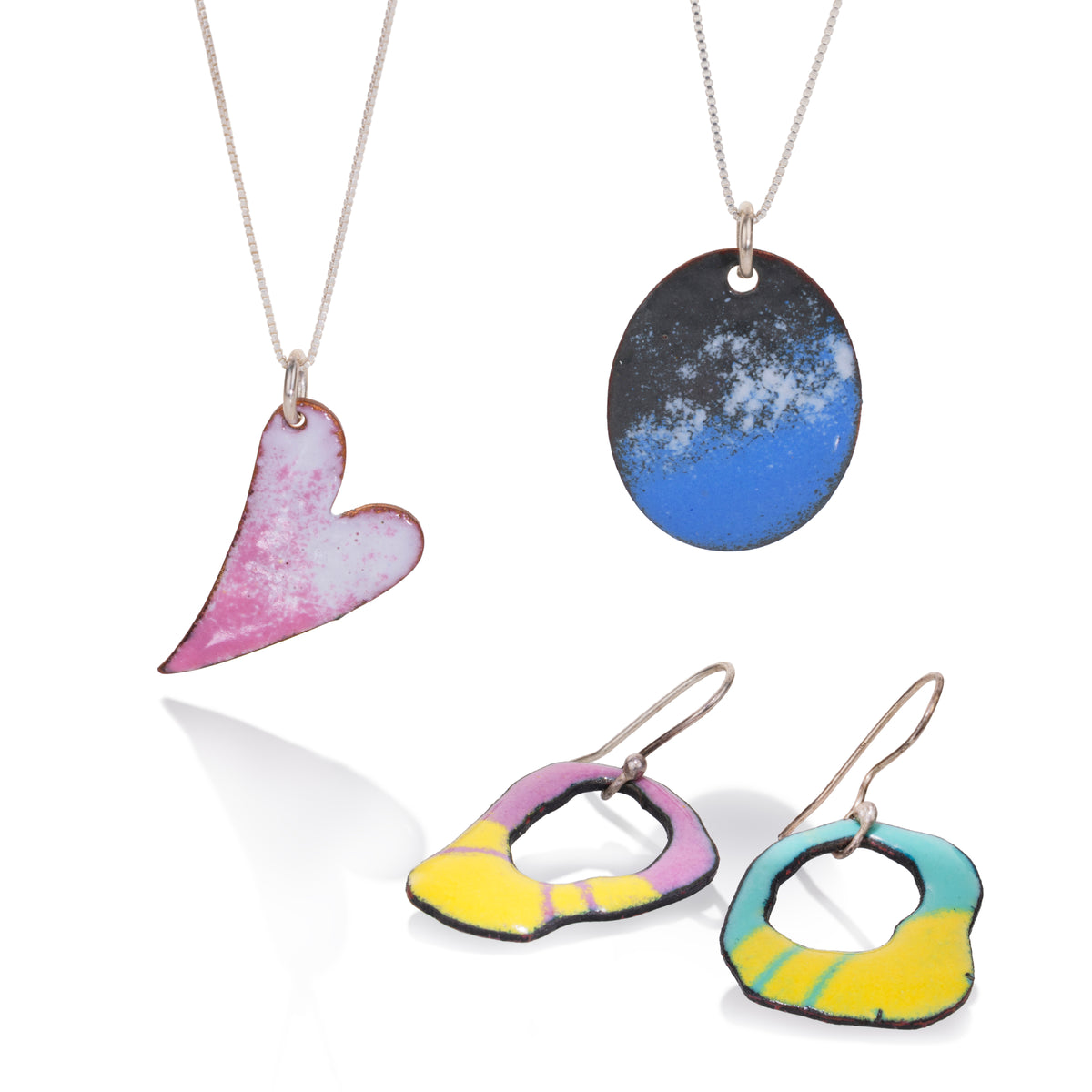 Enameled jewelry featuring a silver necklace with a pink and white heart-shaped pendant, another silver necklace with an oval blue and black pendant, and a pair of matching irregular ring earrings in yellow, pink, and teal with black outlines.