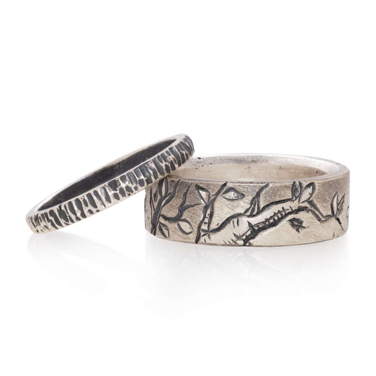 Two silver rings on a white reflective surface, one featuring a textured hammered band and the other engraved with a branch and leaf pattern.