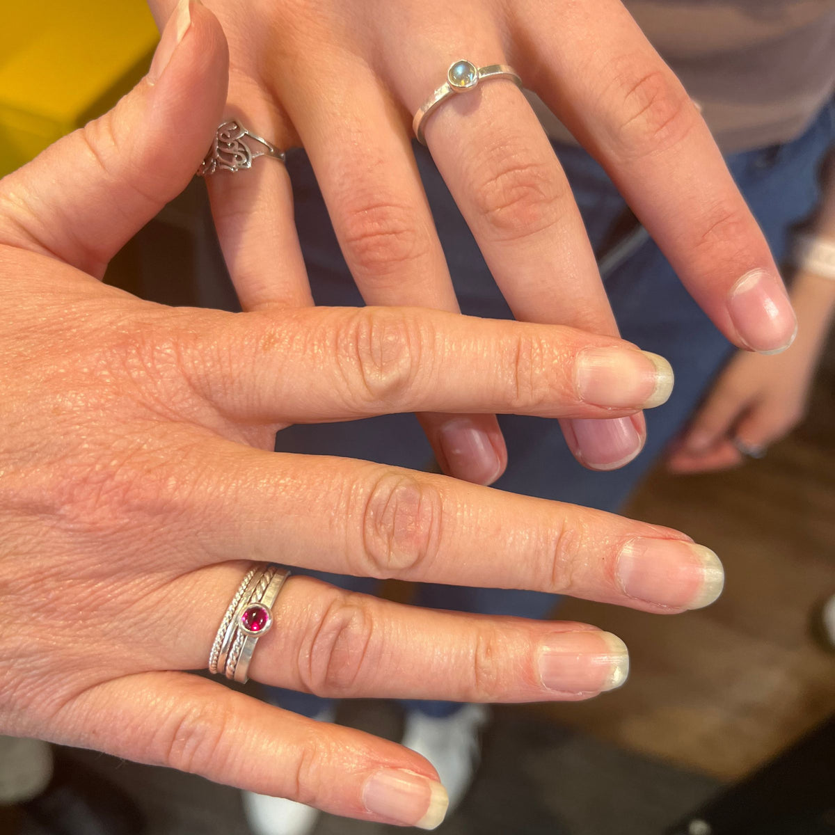 Close-up of hands wearing multiple rings, including a silver band with a round red gemstone and a silver band with a round light blue gemstone, on a person's fingers.